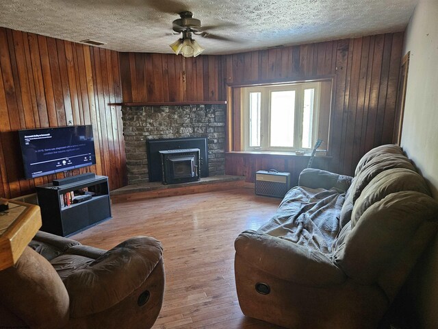 living room with wood-type flooring, ceiling fan, wood walls, a textured ceiling, and a stone fireplace