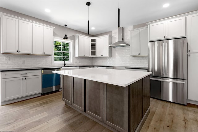 kitchen with hanging light fixtures, wall chimney exhaust hood, light wood-type flooring, white cabinetry, and stainless steel appliances