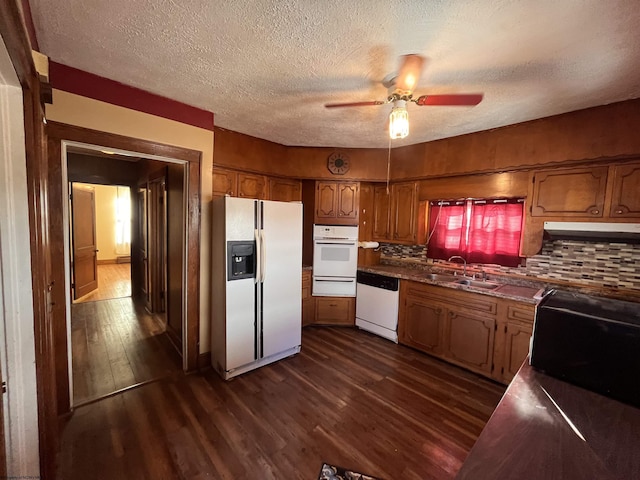 kitchen with white appliances, brown cabinetry, dark countertops, dark wood-style floors, and a sink