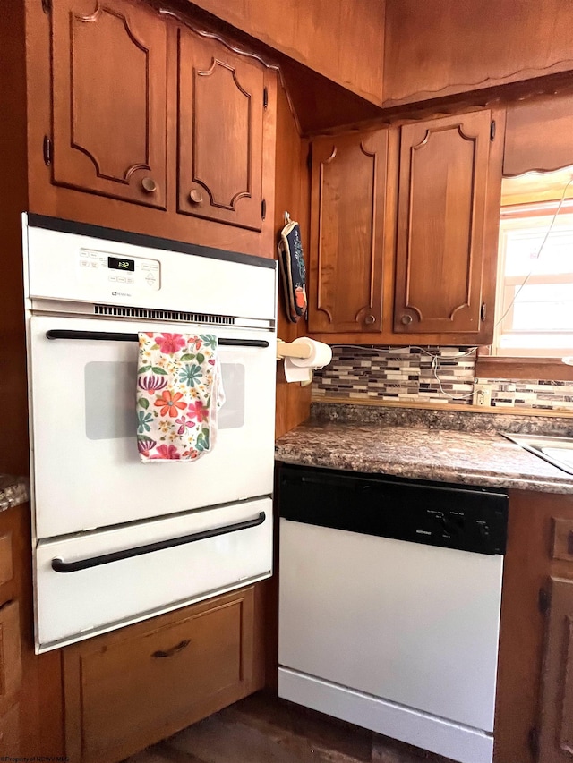 kitchen featuring brown cabinets, a warming drawer, dark countertops, decorative backsplash, and white dishwasher