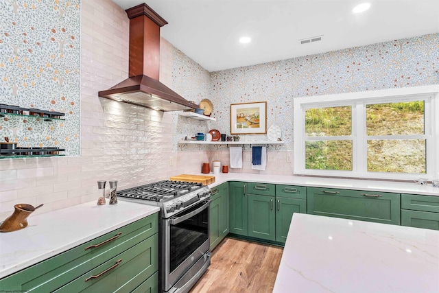 kitchen with light stone counters, stainless steel gas stove, wall chimney exhaust hood, green cabinets, and light hardwood / wood-style floors