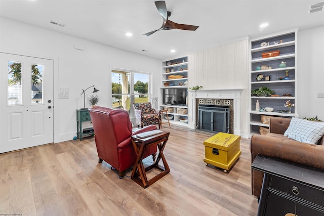 living room with light wood-type flooring, a tiled fireplace, ceiling fan, and built in shelves