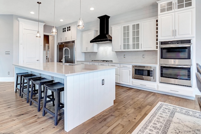 kitchen with wall chimney exhaust hood, a kitchen island with sink, white cabinets, and built in appliances