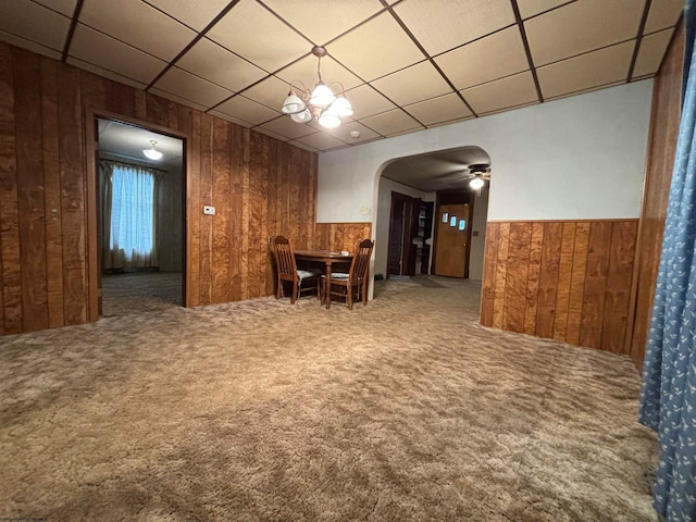 carpeted dining space featuring a paneled ceiling, wooden walls, and ceiling fan with notable chandelier