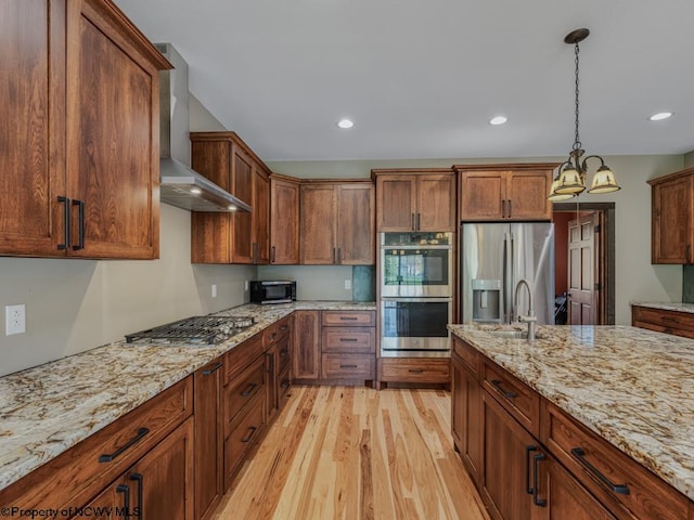 kitchen with appliances with stainless steel finishes, wall chimney exhaust hood, decorative light fixtures, light stone counters, and light hardwood / wood-style flooring