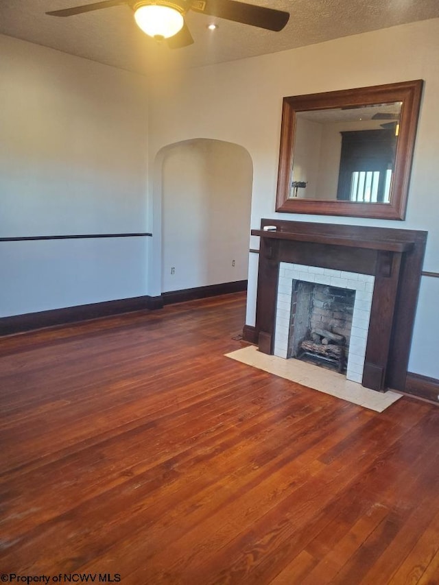 unfurnished living room featuring ceiling fan, a textured ceiling, dark wood-style flooring, baseboards, and a brick fireplace