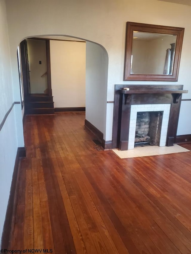 unfurnished living room with arched walkways, a brick fireplace, dark wood-style floors, and baseboards