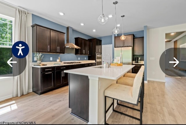 kitchen with light wood-type flooring, a sink, wall chimney range hood, stainless steel appliances, and light countertops