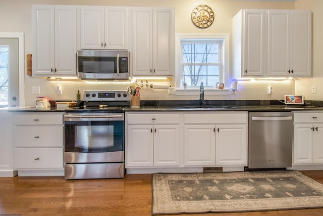 kitchen with appliances with stainless steel finishes, sink, light hardwood / wood-style flooring, dark stone countertops, and white cabinetry