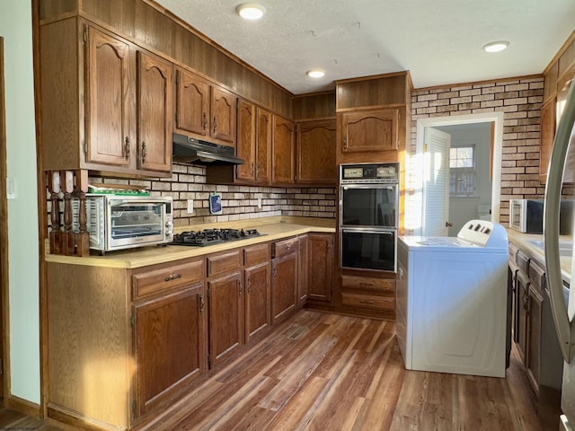 kitchen featuring dark wood-type flooring, black double oven, tasteful backsplash, stainless steel gas cooktop, and washing machine and clothes dryer