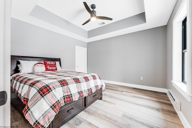 bedroom featuring a baseboard heating unit, ceiling fan, a tray ceiling, and light wood-type flooring