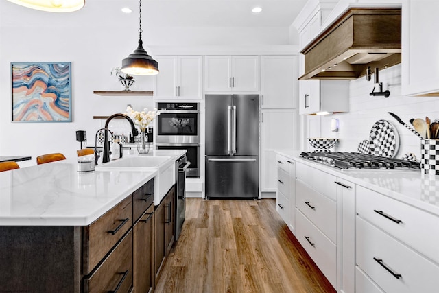 kitchen with custom exhaust hood, appliances with stainless steel finishes, a kitchen island with sink, pendant lighting, and white cabinets