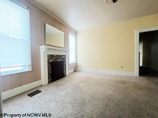 unfurnished living room featuring crown molding, wood walls, a stone fireplace, and carpet flooring