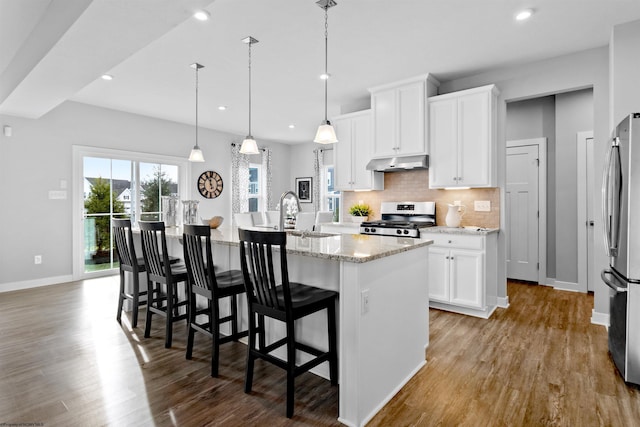 kitchen with stainless steel appliances, light stone countertops, white cabinets, a center island with sink, and decorative light fixtures