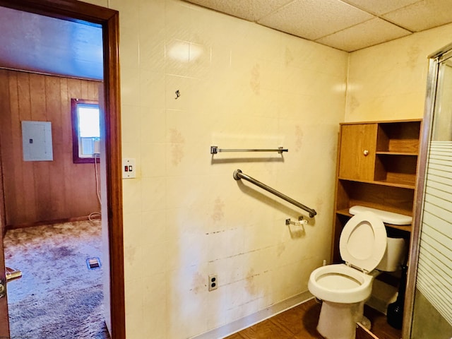 bathroom featuring a paneled ceiling, electric panel, wooden walls, and toilet