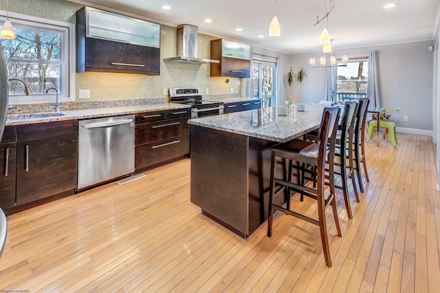 kitchen with a center island with sink, stainless steel appliances, hanging light fixtures, a sink, and wall chimney range hood