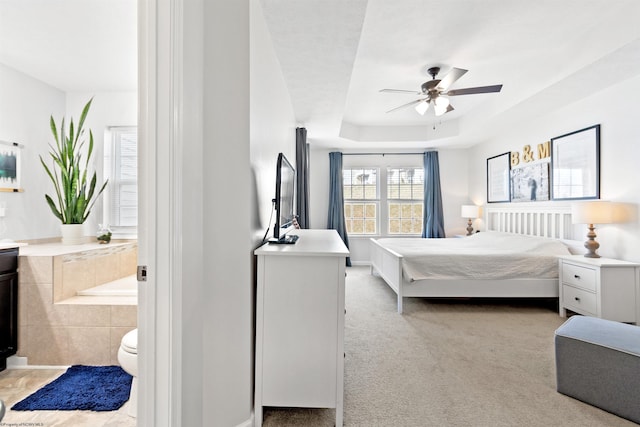 bedroom with a raised ceiling, light carpet, connected bathroom, and ceiling fan