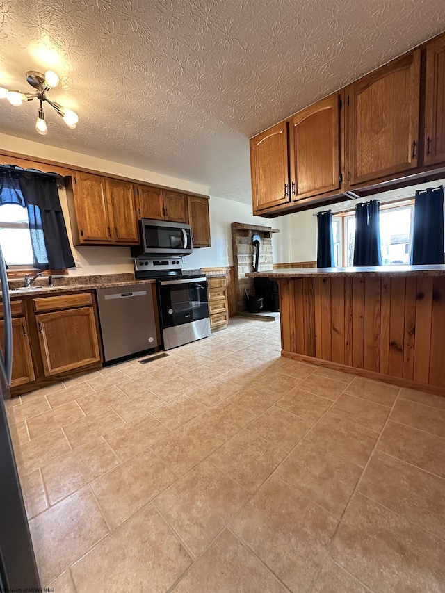 kitchen featuring a textured ceiling, appliances with stainless steel finishes, brown cabinetry, dark countertops, and a wood stove