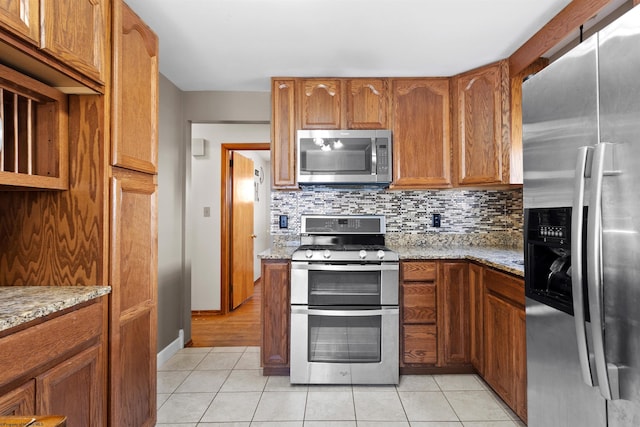 kitchen with light tile patterned floors, decorative backsplash, brown cabinets, light stone countertops, and stainless steel appliances