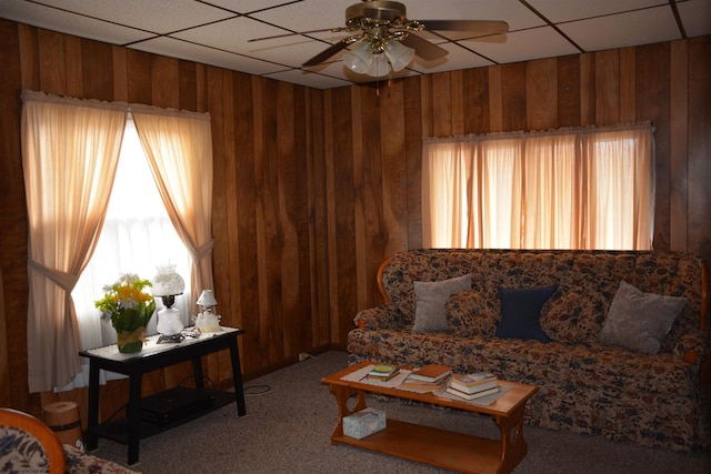 carpeted living room with wooden walls, a ceiling fan, and a drop ceiling