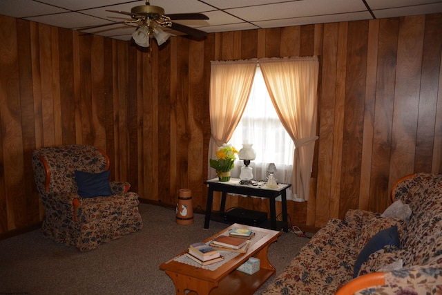 living area with a paneled ceiling, wood walls, carpet floors, and a ceiling fan