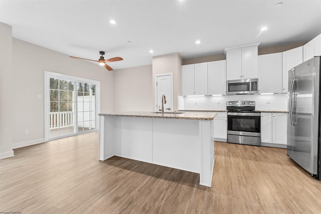 kitchen with light wood finished floors, a center island with sink, appliances with stainless steel finishes, light stone countertops, and a sink