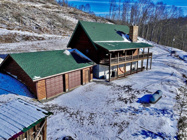 snow covered property with driveway, a garage, log veneer siding, a balcony, and a chimney