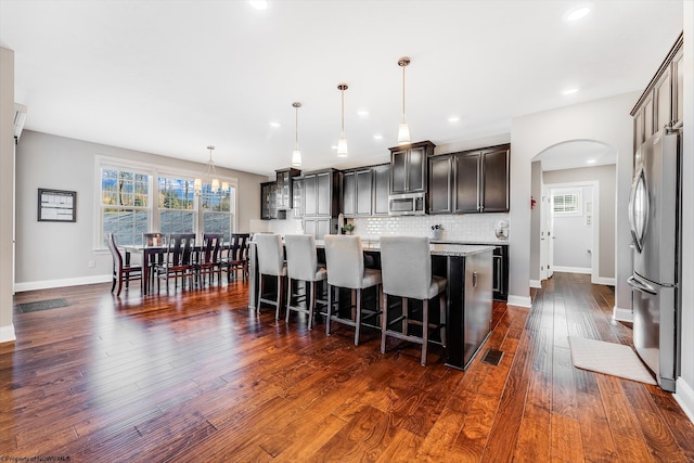 kitchen with arched walkways, dark wood finished floors, appliances with stainless steel finishes, tasteful backsplash, and a kitchen bar