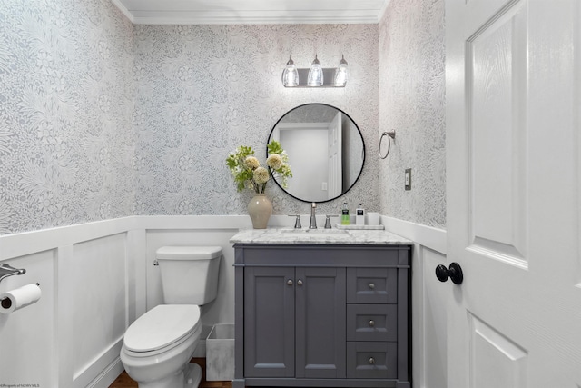 half bath with toilet, a wainscoted wall, vanity, ornamental molding, and wallpapered walls