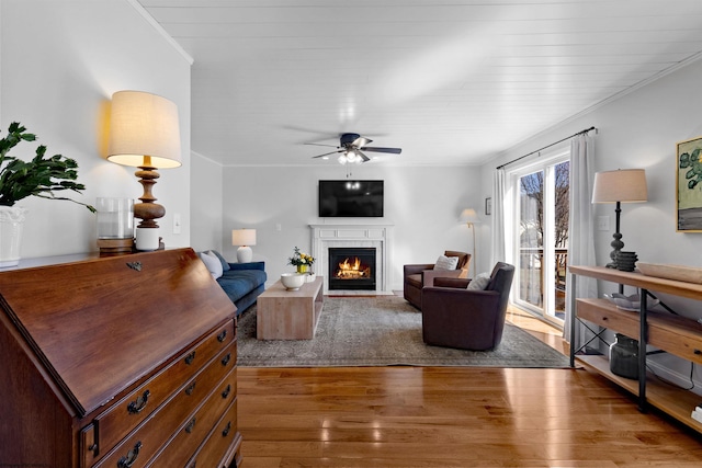 living area featuring a warm lit fireplace, ornamental molding, wood finished floors, and a ceiling fan