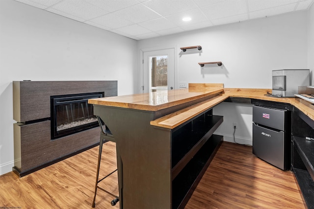 kitchen featuring a drop ceiling, a breakfast bar, wood finished floors, wooden counters, and a multi sided fireplace