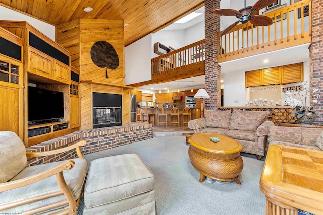 carpeted living room featuring a towering ceiling, a ceiling fan, a glass covered fireplace, wood walls, and wooden ceiling