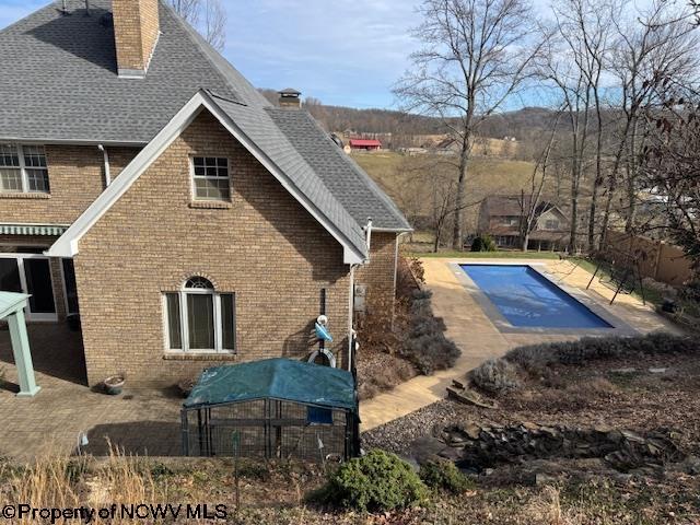 view of side of home featuring an outdoor pool, a chimney, roof with shingles, a patio area, and brick siding