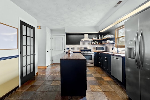 kitchen featuring stone tile floors, wall chimney exhaust hood, stainless steel appliances, dark cabinetry, and a sink