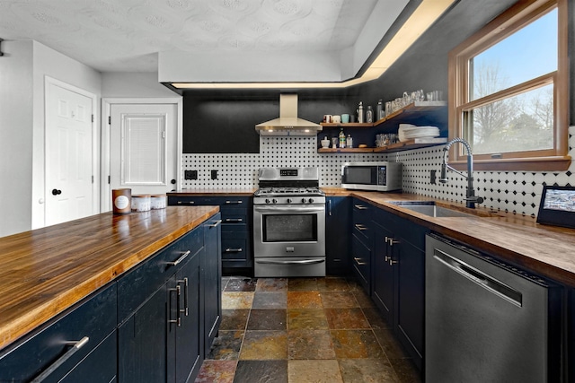 kitchen with tasteful backsplash, appliances with stainless steel finishes, a sink, wood counters, and wall chimney range hood