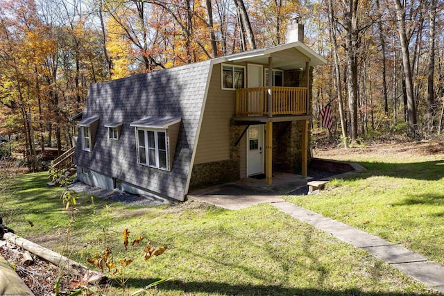 view of front facade featuring a shingled roof, a gambrel roof, a chimney, a front yard, and a wooded view