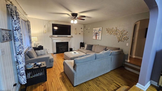 living room featuring a ceiling fan, baseboards, a fireplace with flush hearth, arched walkways, and wood-type flooring