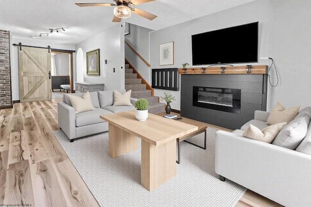 living room with stairway, a barn door, light wood-style floors, a glass covered fireplace, and a ceiling fan