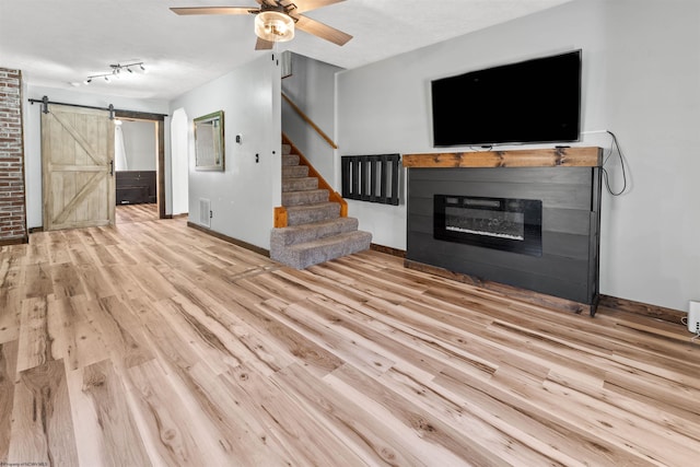 unfurnished living room featuring a glass covered fireplace, wood finished floors, a barn door, stairway, and ceiling fan