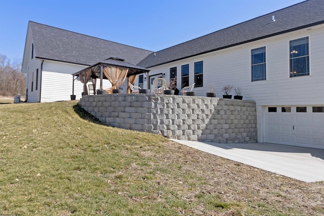 rear view of property featuring a lawn, a gazebo, roof with shingles, concrete driveway, and a garage