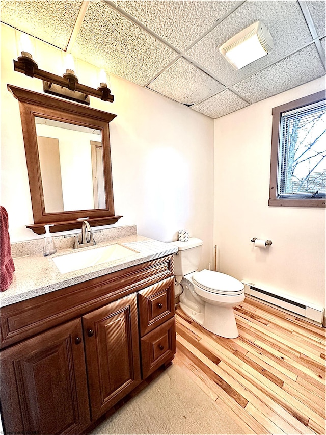 bathroom with vanity, wood finished floors, a baseboard radiator, a paneled ceiling, and toilet