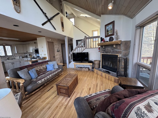 living room with stairs, light wood-type flooring, a stone fireplace, wooden ceiling, and high vaulted ceiling