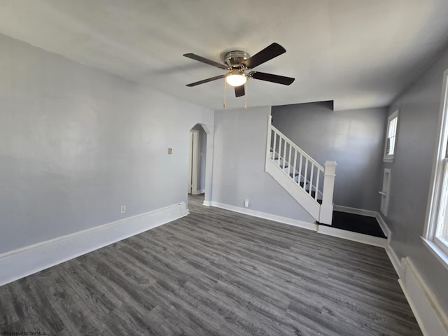 unfurnished living room featuring baseboards, ceiling fan, stairs, arched walkways, and dark wood-style flooring