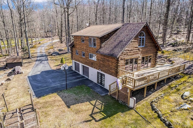 exterior space featuring a forest view, a shingled roof, log siding, a deck, and driveway