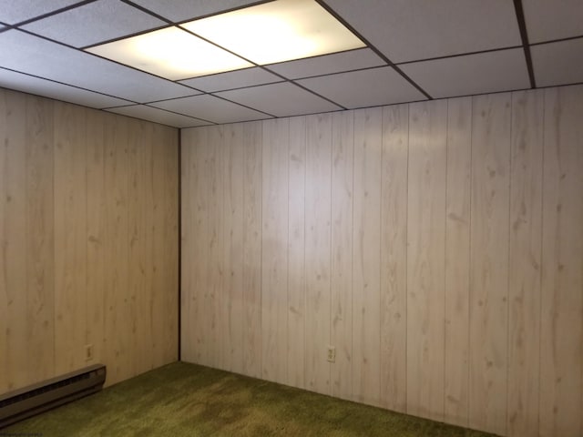 carpeted empty room featuring a drop ceiling, baseboard heating, and wood walls