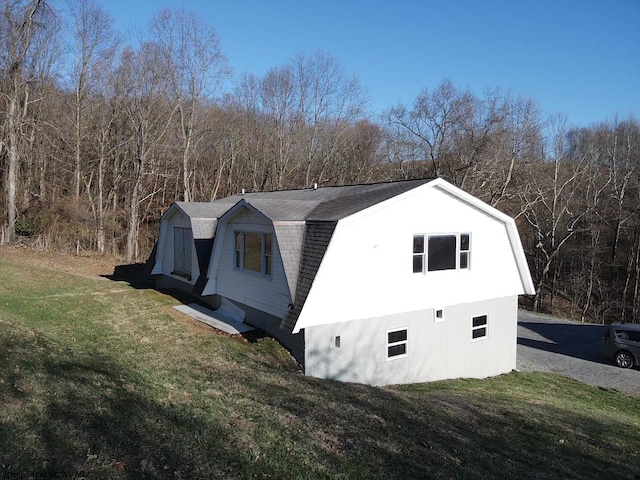 view of side of property with a gambrel roof, a view of trees, a lawn, and a shingled roof