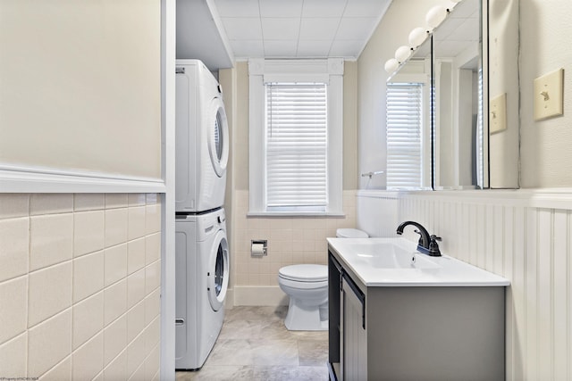 bathroom with vanity, stacked washer / dryer, tile walls, a wainscoted wall, and toilet