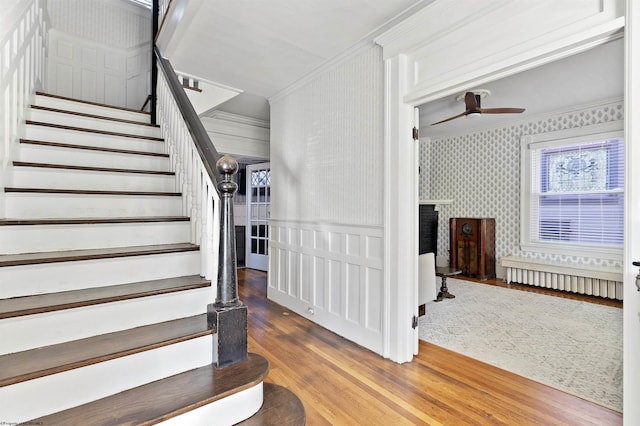 stairway with wallpapered walls, a ceiling fan, wood finished floors, a fireplace, and ornamental molding