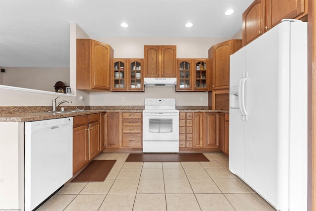 kitchen featuring a sink, under cabinet range hood, glass insert cabinets, brown cabinetry, and white appliances