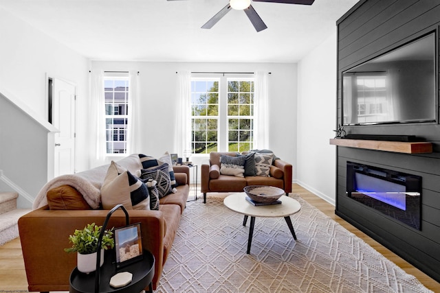 living area featuring stairway, baseboards, a glass covered fireplace, light wood finished floors, and a ceiling fan